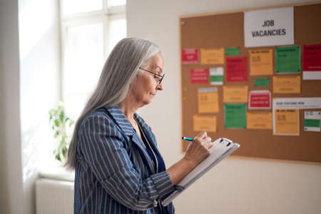 Job center employee with file form standing in front of employment noticeboard.