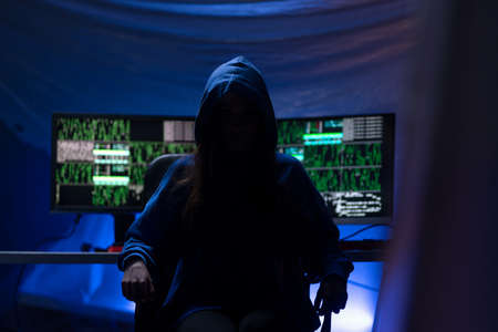 Hooded anonymous hacker by computer in the dark room at night, cyberwar concept.