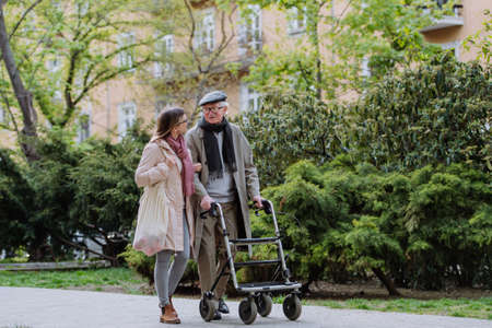 Senior man with walking frame and adult daughter outdoors on a walk in park.の写真素材