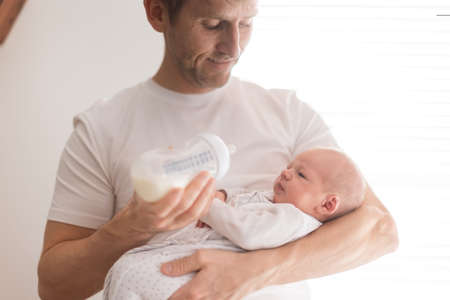 Father holding and feeding his newborn son with milk bottle at home.