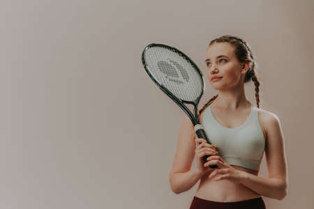 Young sporty woman in sports clothes holding tennis racket on her shoulder while standing against...