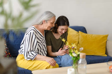 Happy senior grandmother with teenage granddaguhter sitting on sofa and using smartphone together at...