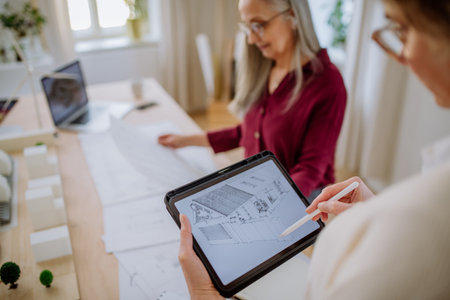 Mature women eco architects with blueprints working on tablet together in office.