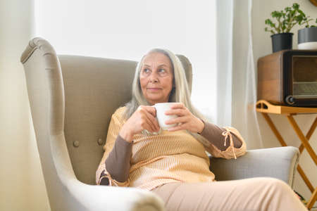 Relaxed senior woman with cup of tea sitting in armchair at home.