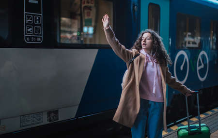 Young traveler woman with luggage standing in platform and saying goodbye, waveing somebody in trainの写真素材