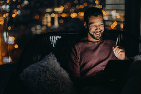 Young African AMerican man sitting on balcony with urban view and using tablet at nightの写真素材