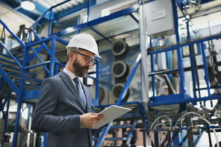 Chief Engineer in the hard hat walks through industrial factory while holding tablet.