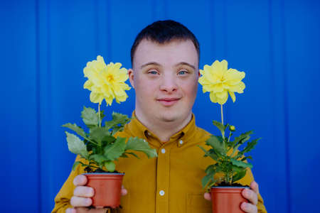 Happy young man with Down syndrome looking at camera and holding pot flowers against blue background.の写真素材