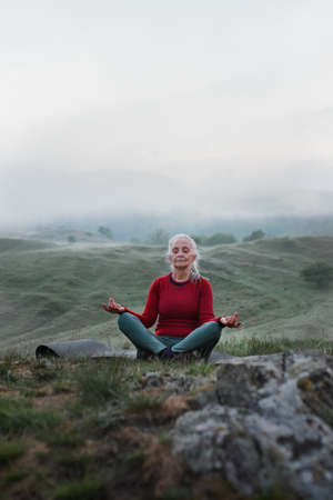 Senior woman doing breathing exercise in nature on early morning with fog and mountains in background.の写真素材