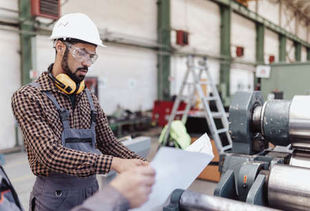 Heavy industry worker with safety headphones and hard hat in industrial factory holding blueprintsの写真素材