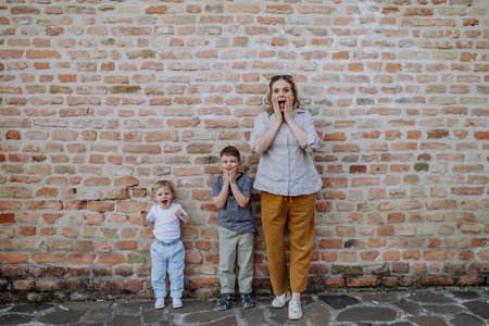 Young mother with her little children standing by brick wall and making funny faces in street summer.の写真素材