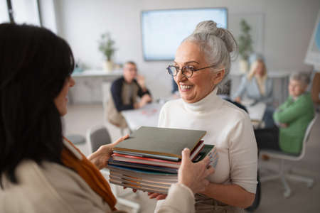 Happy senior woman student giving books to teacher in classroom.の写真素材