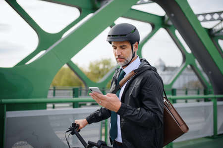 Businessman commuter on the way to work, pushing bike on bridge and texting on mobile phone,...