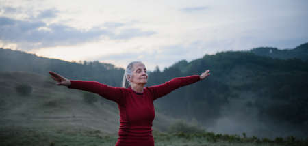 Senior woman doing breathing exercise in nature on early morning with fog and mountains in...