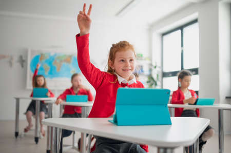 Happy schoolgirl with hands up using digital tablet during lesson in classroom at primary school.