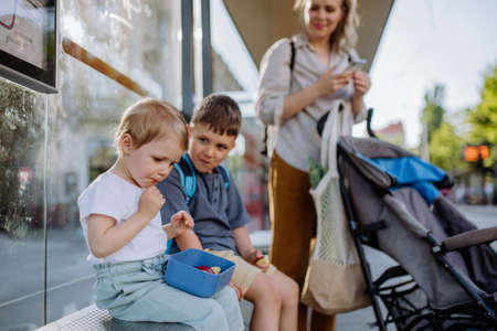 Young mother with little kids waiting on bus stop in city and eating fruit snack.の写真素材