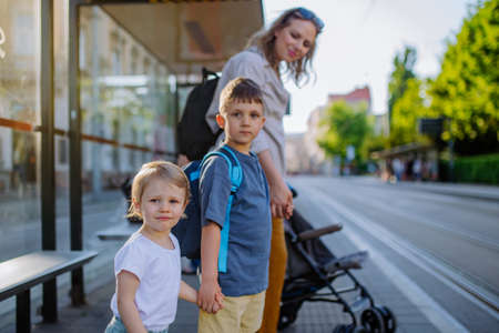 Young mother commuter with little kids on the way to school, waiting for bus in city.の写真素材