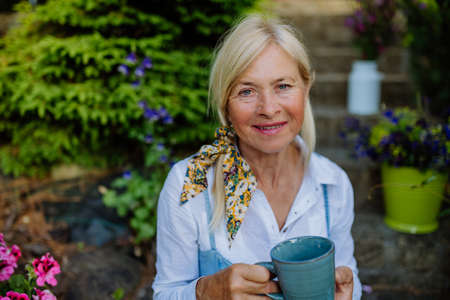 Portrait of senior woman with coffee sitting on terrace in summer, resting and looking at camera.の写真素材