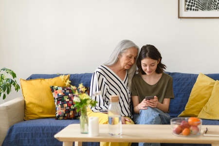 Happy senior grandmother with teenage granddaguhter sitting on sofa and using smartphone together at...