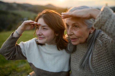 Portrait of happy senior grandmother with teenage granddaguhter sitting on grass and looking into the distance in nature on spring day during sunset.の写真素材