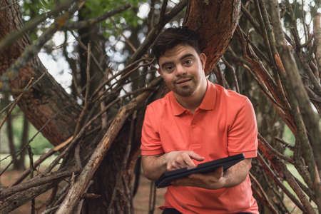 Portrait of happy young man with down syndrome standing outdoors in park, using digital tablet.