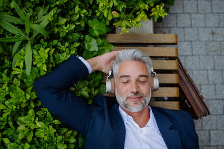Top view of mature businessman enjoying music in headphones relaxing on bench in city park during break at work.の写真素材
