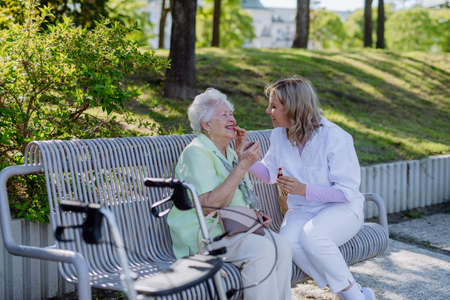 Caregiver helping senior woman to apply lipstick when sitting on bench in park in summer.の写真素材