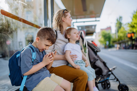 Young mother with little kids waiting on bus stop in city, scrolling on mobile phone.の写真素材