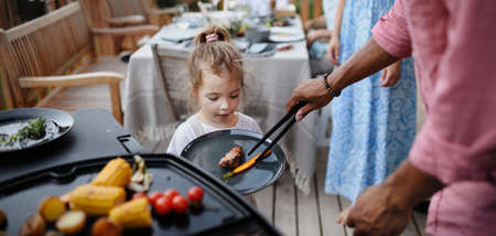 Father putting grilled meat and vegetable on plate tohis daughter during family summer garden party.の写真素材
