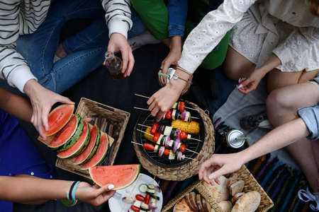 A close-up of young friends putting corn and skewers on grill and having barbecue when camping in...