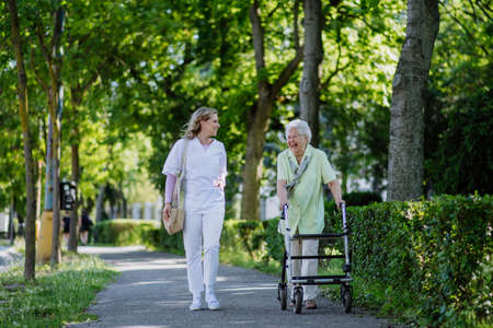 Caregiver with senior woman on walk with walker in park with shopping bag.の写真素材