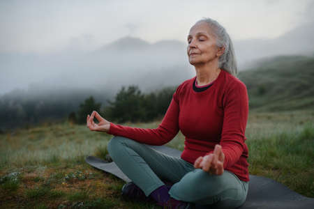 Senior woman doing breathing exercise in nature on early morning with fog and mountains in background.の写真素材