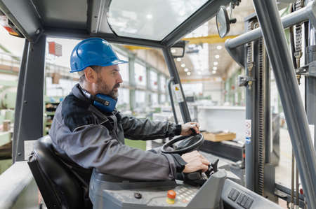 Happy mature man fork lift truck driver lifting pallet in storage warehouse .