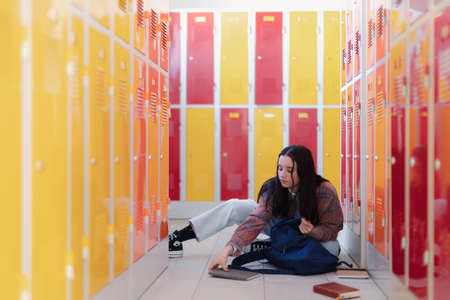 Teenage student sitting in corridor near colorful lockers adn packing book to backpack in campus hallway, back to school concept.の写真素材