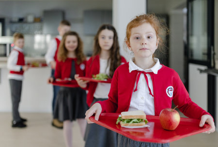 Happy schoolchildren in uniforms holding trays with lunch and standing in queue in school canteen.