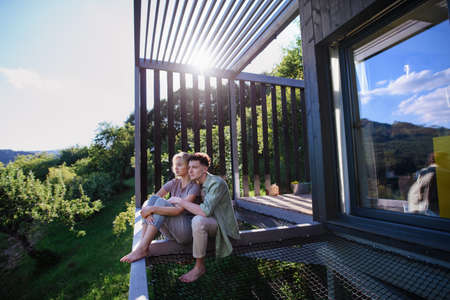 Young couple sitting and cuddling in hammock terrace in their new home in tiny house in woods, sustainable living concept.の写真素材