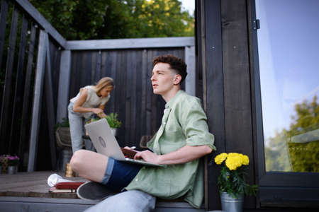 Happy young couple with laptop resting outdoors in a tiny house, weekend away and remote office...