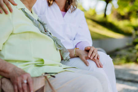 Close-up of caregiver consoling senior woman and touching her hand when sitting on bench in park in summer.の写真素材