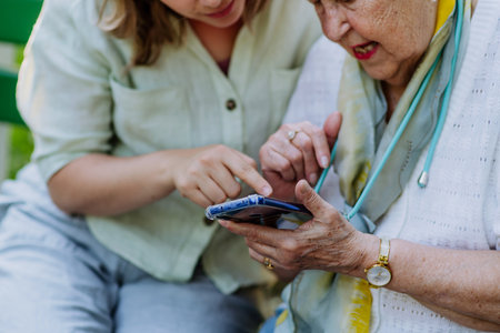 Adult granddaguhter helping her grandmother to use cellphone when sitting on bench in park in summer.の写真素材
