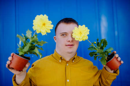 Happy young man with Down syndrome looking at camera and holding pot flowers against blue background.の写真素材
