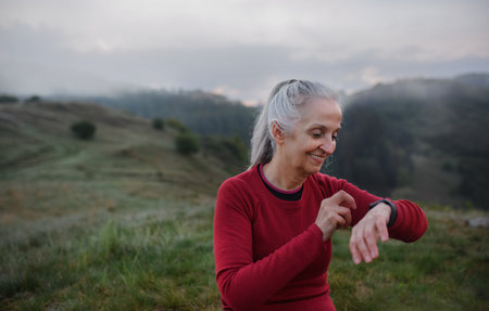 Senior woman jogger setting and looking at sports smartwatch, checking her performance in nature on early morning with fog and mountains in background.の写真素材