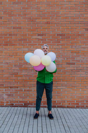 Fun portrait of happy energetic mature man holding balloons in street and hiding behind them, feeling free.の写真素材