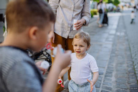 Little boy blowing bubbles with his little sister in city street in summer.