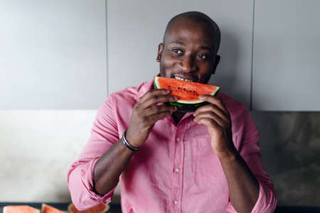 Multiracial man eating watermelon in his kitchen during hot sunny days.の写真素材