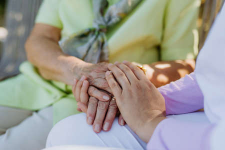 Close-up of caregiver consoling senior woman and touching her hand when sitting on bench in park in...