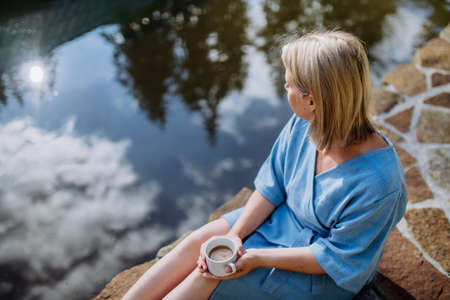 Happy young woman sitting by pond near cottege and enjoying cup of morning coffee on summer vacation in mountains.の写真素材