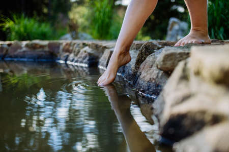 Unrecognizable young woman is dipping her foot in cool water of pond, refreshing and hardening concept.の写真素材