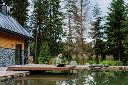 Woman, freelancer working on laptop, sitting on a pier by the backyard lake, a concept of remote office, work during vacation.の写真素材
