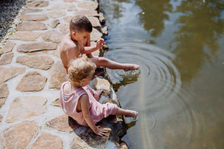 Cute girl and boy sitting together on the footpath by the lake, dangle their feet in the water,...
