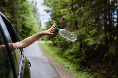 Womans hand throwing away plastic bottle from car window on the road in green nature, environmental...